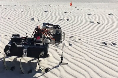 A small black and silver legged robot with red wiring in white sand